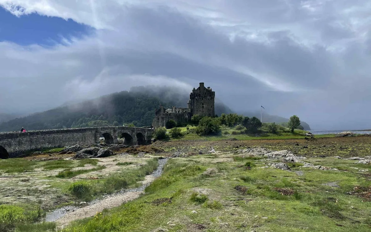 Eilean Donan Castle and its arched bridge seen from a grassy shore, with low cloud on the hill behind