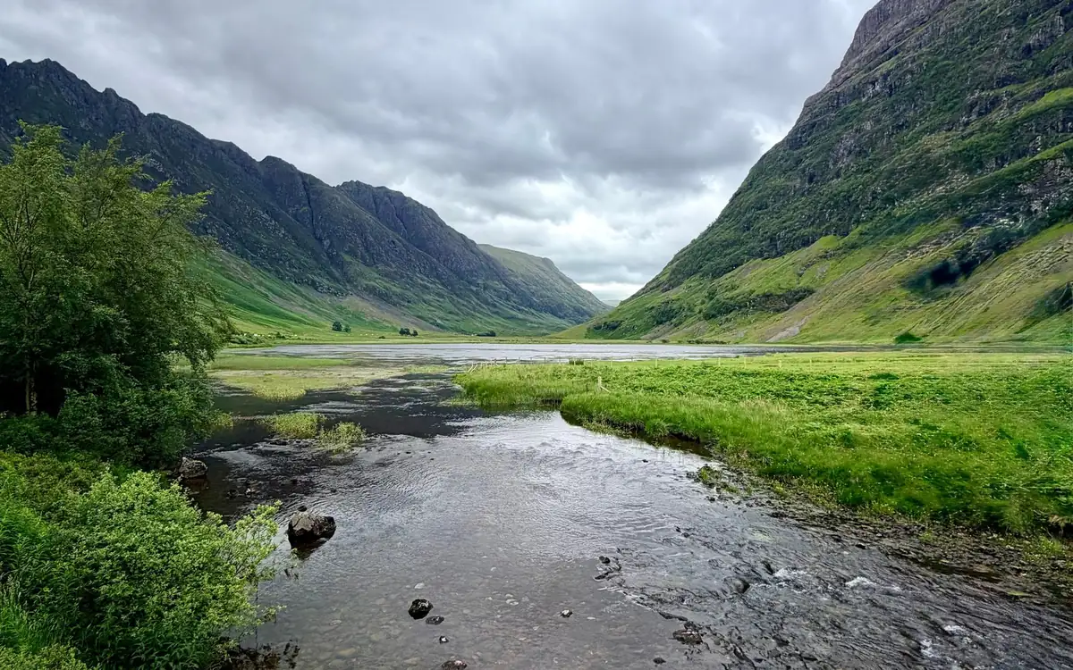Un río poco profundo que desemboca en un lago entre laderas verdes escarpadas y crestas oscuras