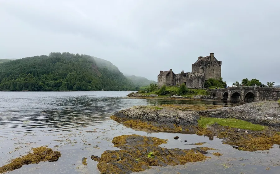 Eilean Donan Castle oltre il loch, con rocce ricoperte di alghe e nuvole basse sulle colline