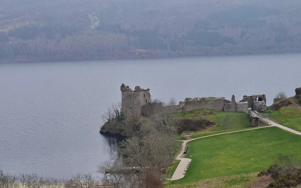 Ein verfallenes Tower House und Burgmauern auf einer grasbewachsenen Landzunge über einem weiten grauen Loch, vom Hang aus gesehen